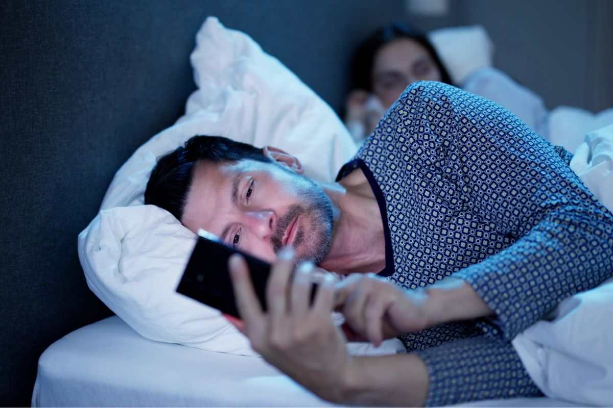 uomo a letto con telefono in mano e donna alle spalle